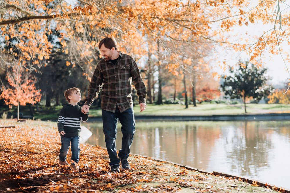 Father and young son walk hand-in-hand along a pond in a park surrounded by vibrant autumn foliage.