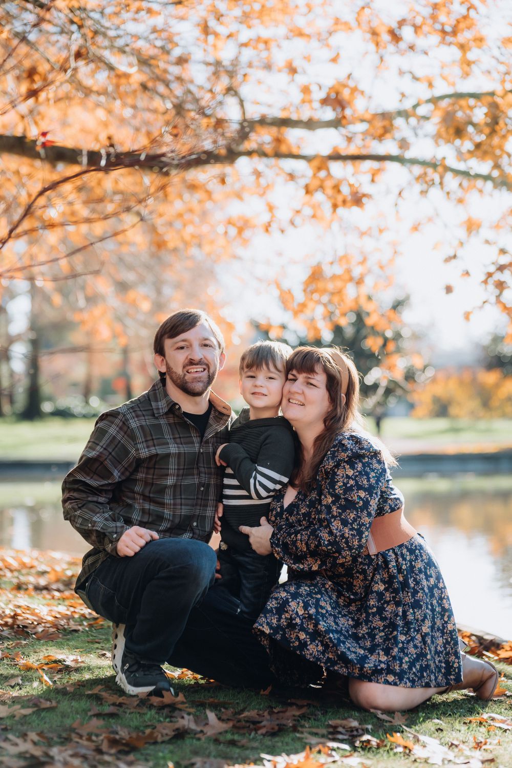 Family of three posing outdoors in autumn park with golden foliage and pond backdrop on sunny fall day.