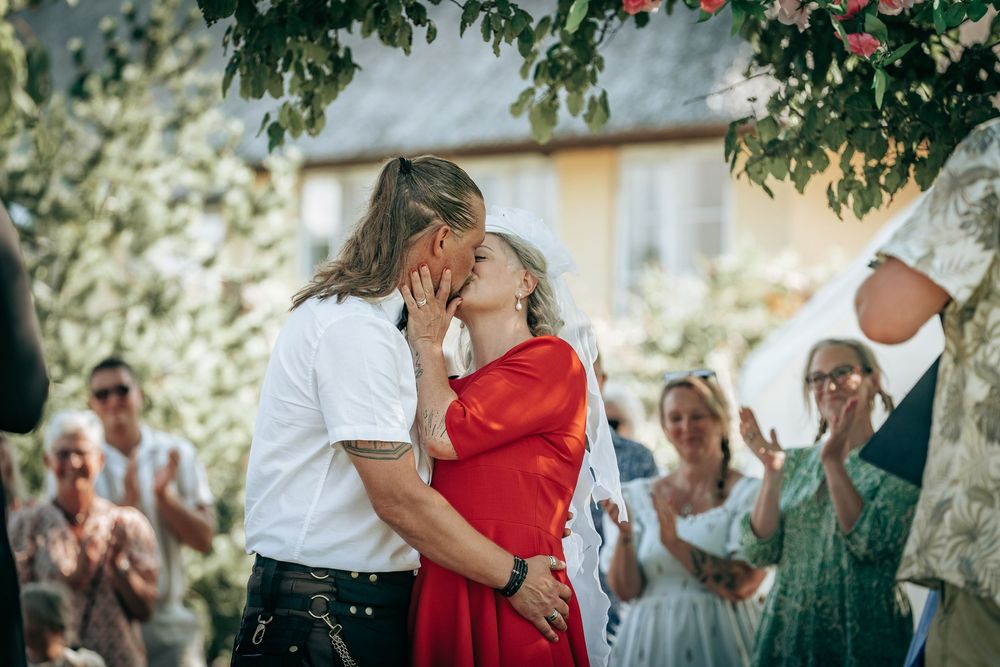 Kys mellem brudepar under udendørs bryllup med gæster – romantisk øjeblik fanget af fotograf i Jægerspris, Frederikssund og Nordsjælland