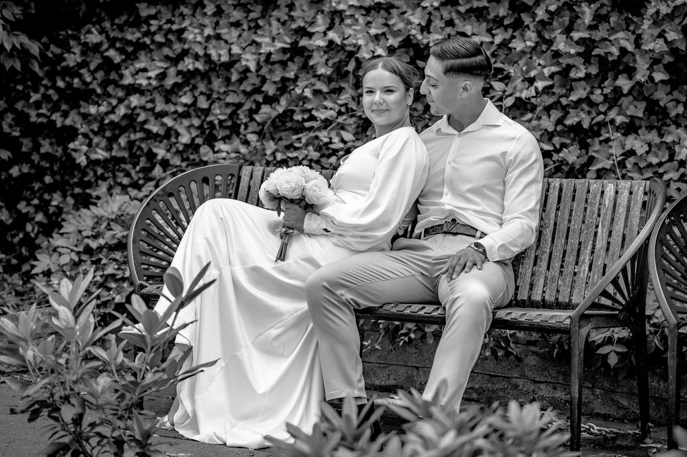 Romantisk bryllupsfoto af et brudepar, der sidder tæt på en bænk omgivet af grønne planter – fotograferet af CHH_foto i Jægerspris, Frederik