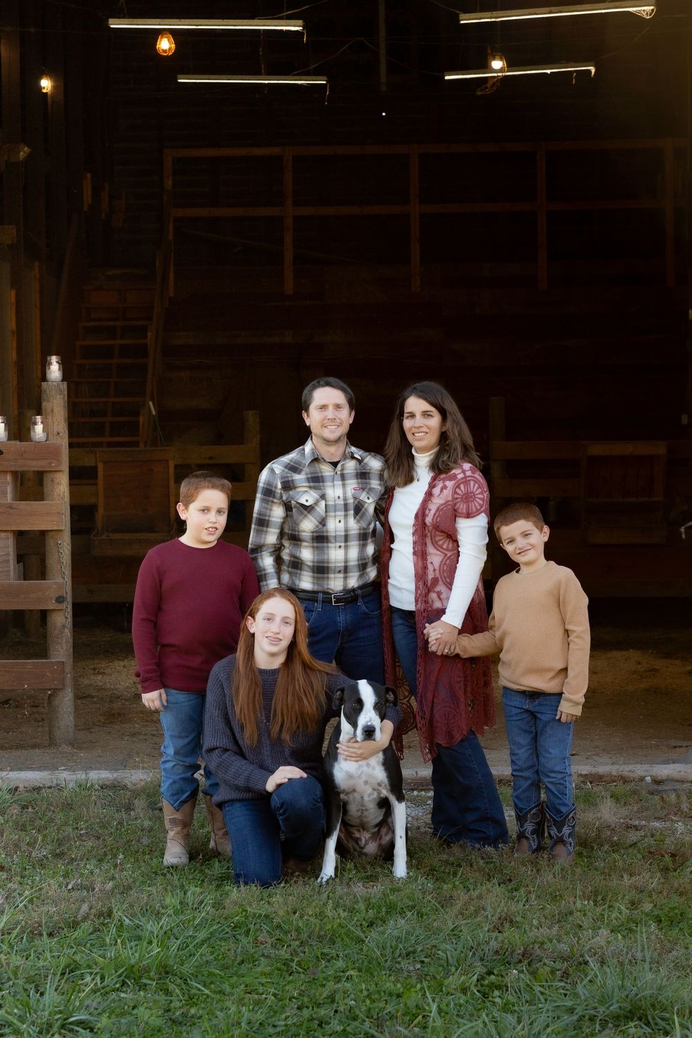 Family gathers with their dog for outdoor portrait session in front of rustic barn setting.