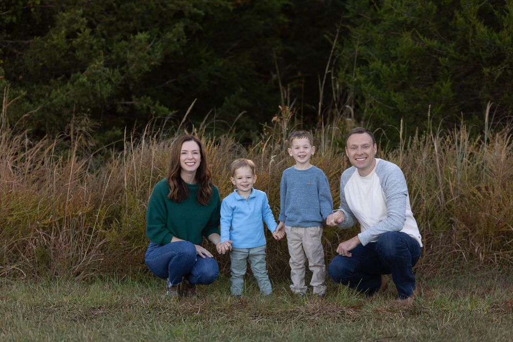 A family group poses together against tall brown grass and trees in an outdoor setting.