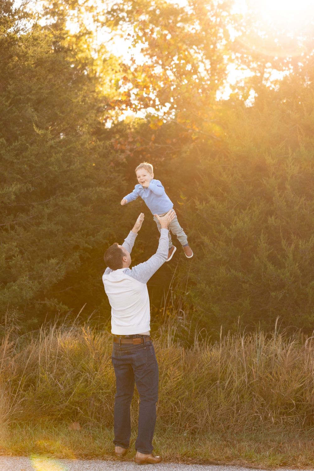 A person in a white top lifts a small child playfully into the air during golden hour at sunset.