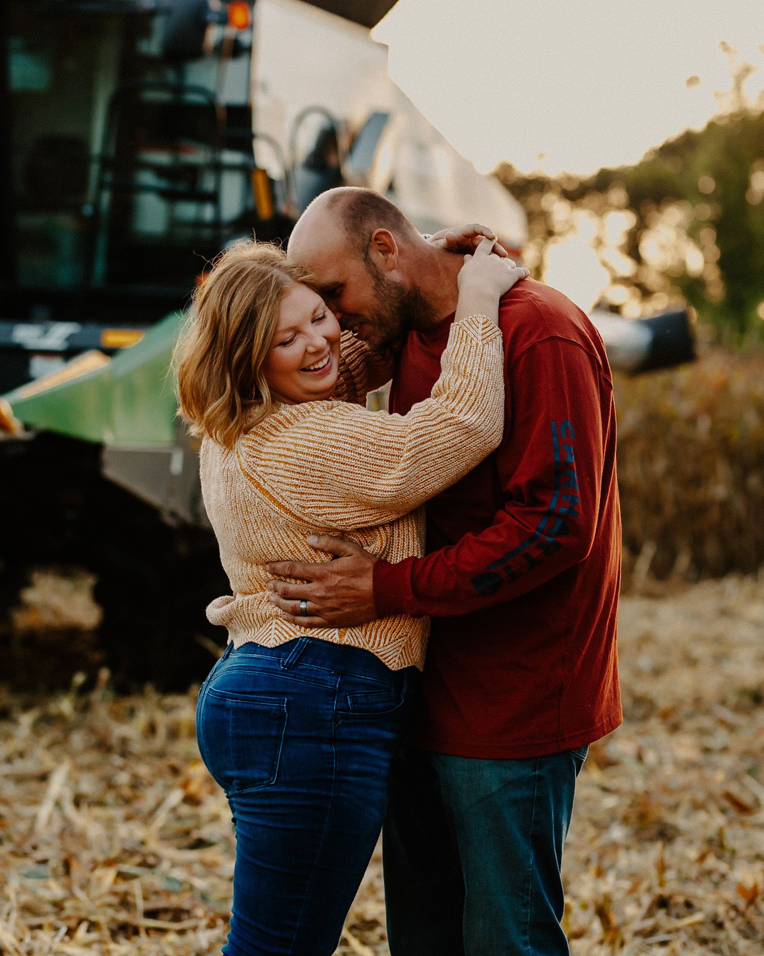 Laughing couple embracing warmly at golden hour in a harvested farm field with equipment behind them.