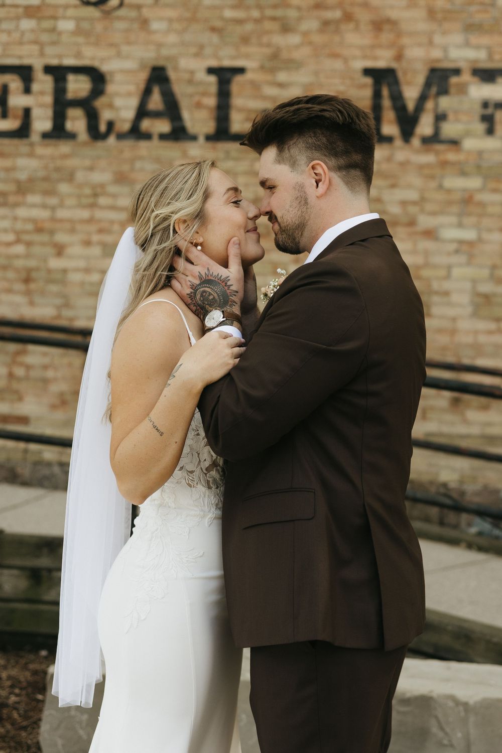 A romantic wedding moment with a bride in white dress and groom in dark suit sharing a tender kiss against brick wall.