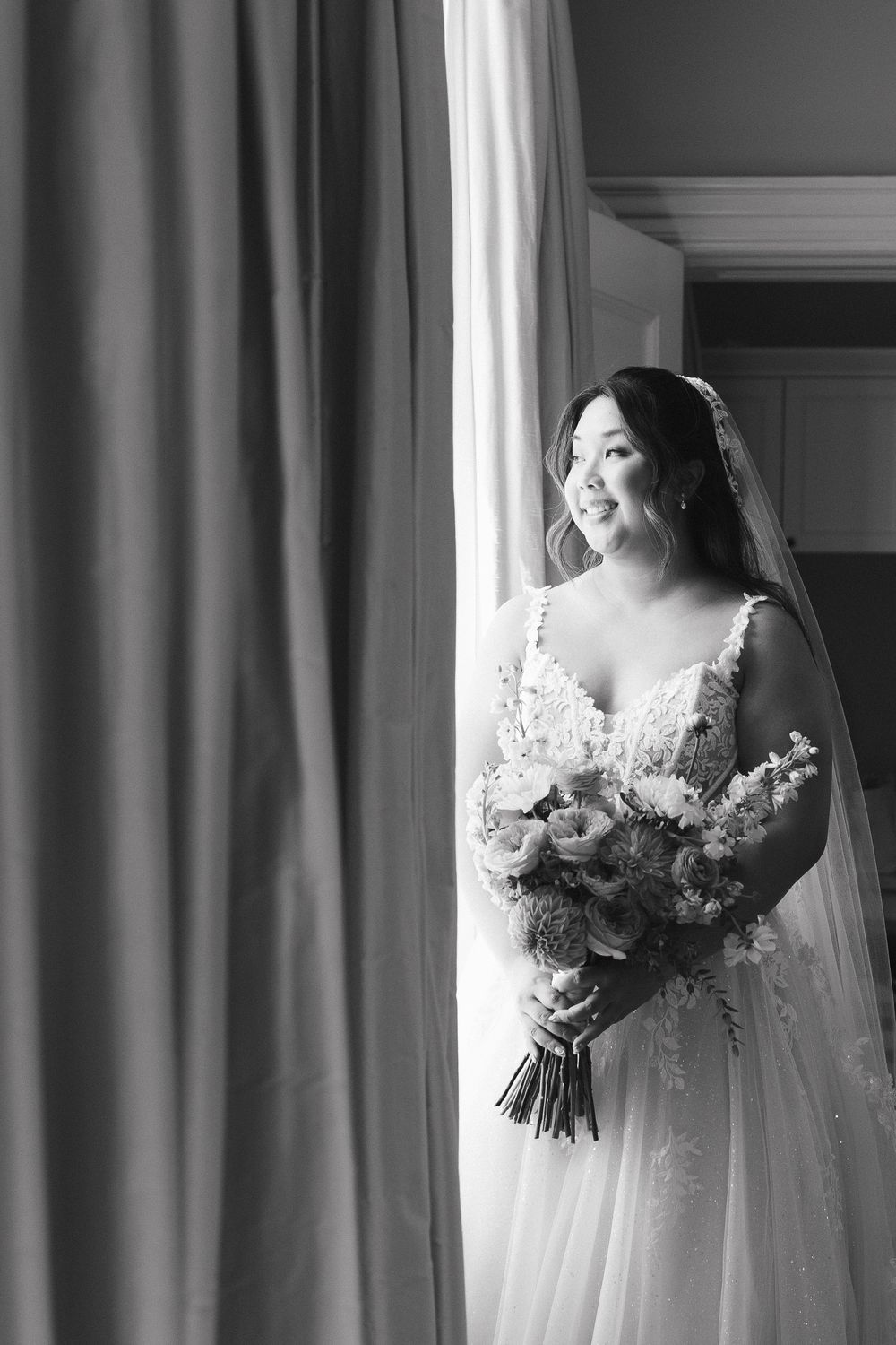 Black and white wedding photo of a bride in a lace gown holding a floral bouquet while standing by a window at Langdon Hall
