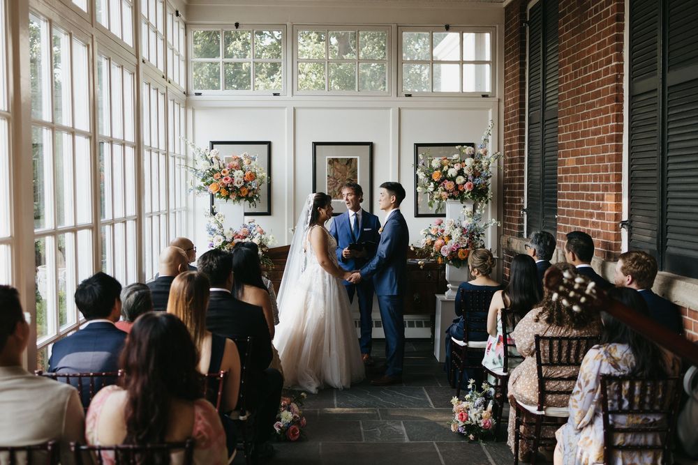 Wedding guests gather in a bright sunlit room with exposed brick walls and large windows during an indoor ceremony at Langdon Hall