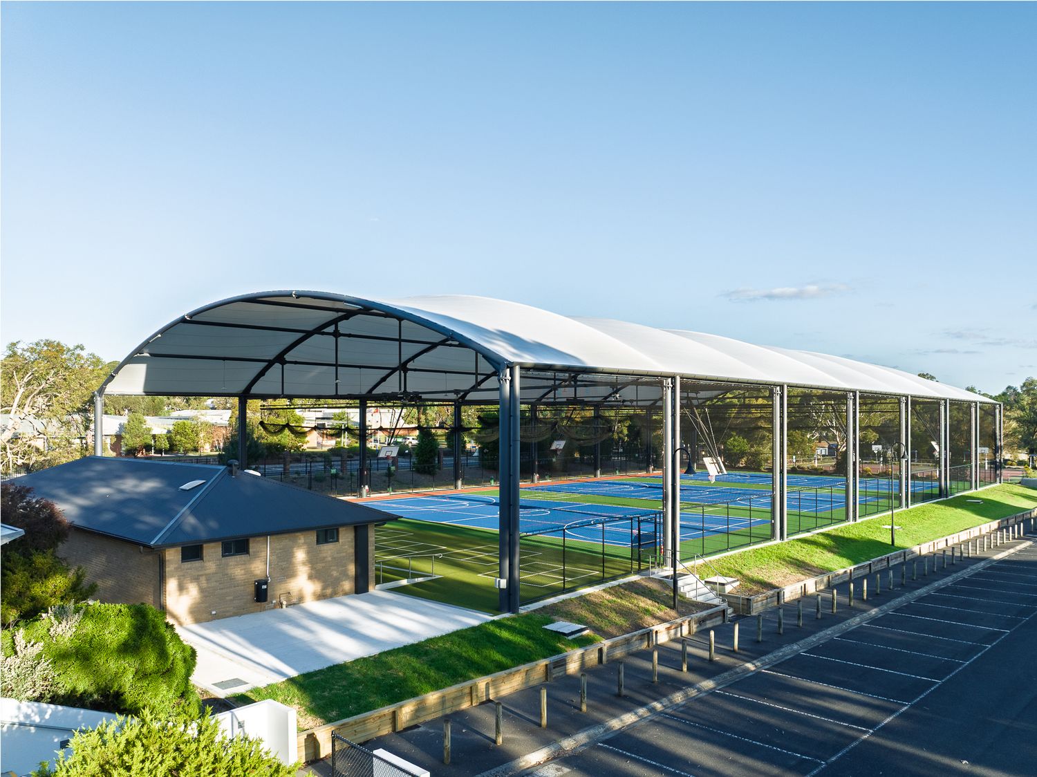 Modern covered tennis courts facility with arched roof structure, blue courts, and adjacent parking area.