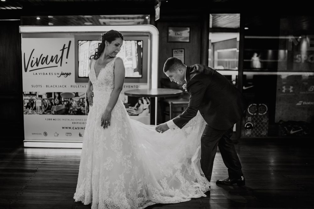 Groom adjusting bride's lace wedding dress train in black and white photo at elegant venue. Fotografía de Bodas en Formosa