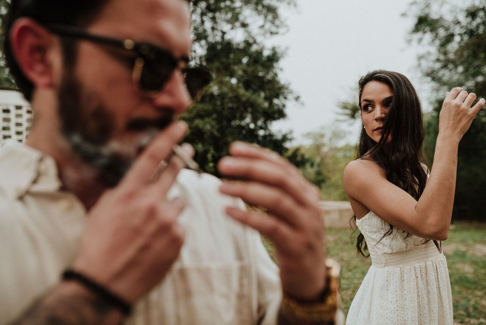 Man in sunglasses and beige shirt in foreground, woman in white dress adjusting hair in background outdoors. Fotógrafo en Formosa