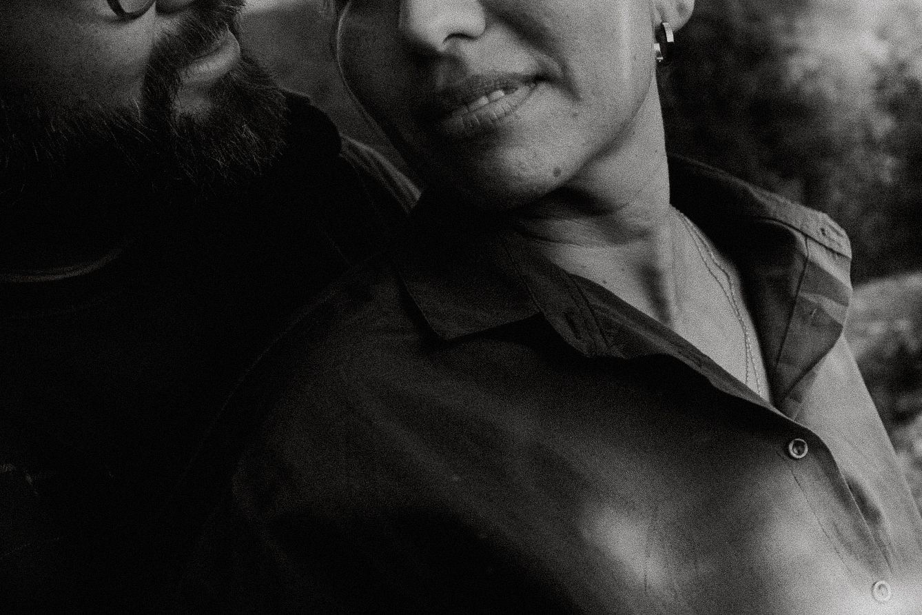 Black and white close-up portrait of a couple embracing outdoors, man with beard nuzzling woman wearing a dark shirt.