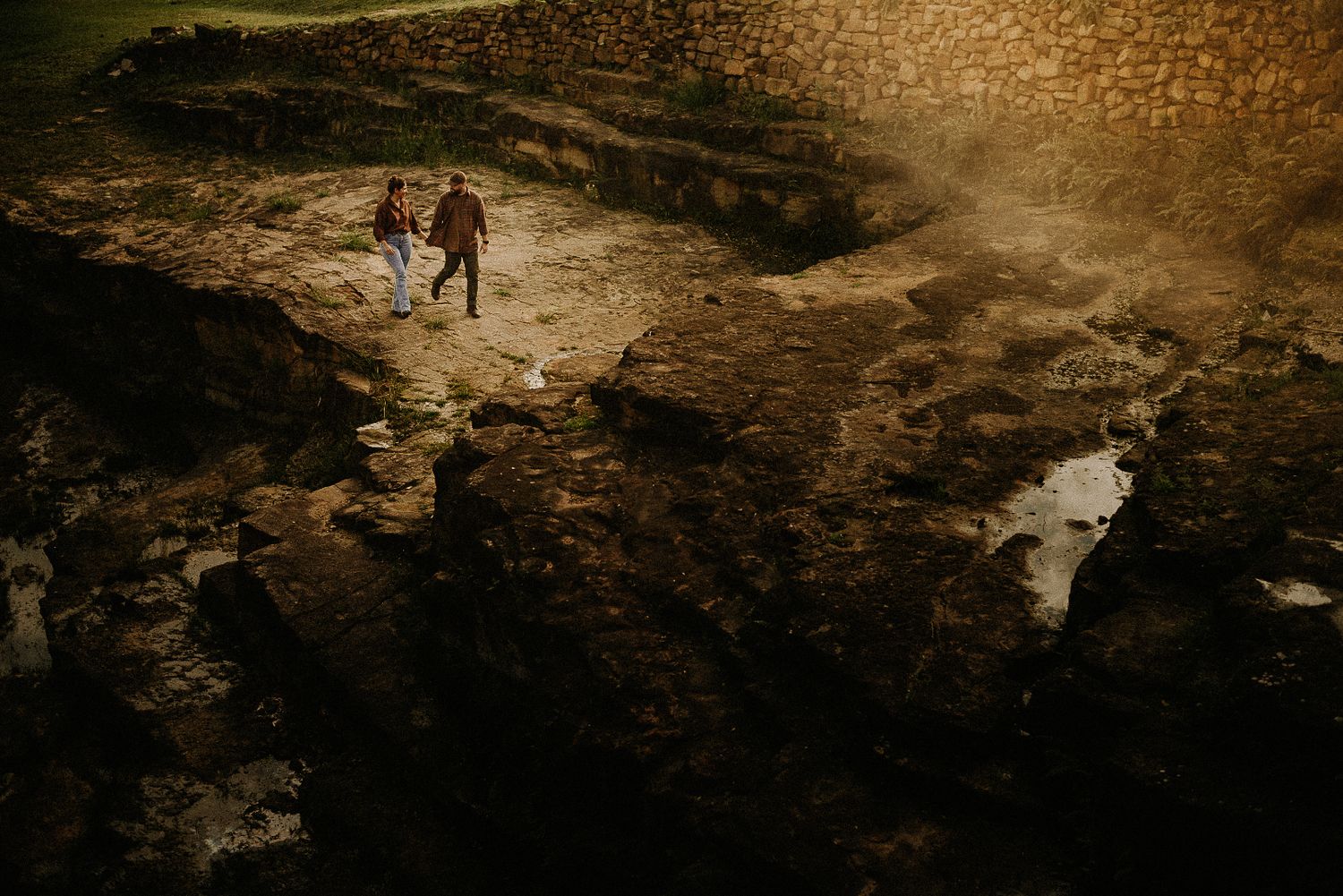 Two people walk hand-in-hand across dramatic rocky terrain at night, illuminated by warm golden light.