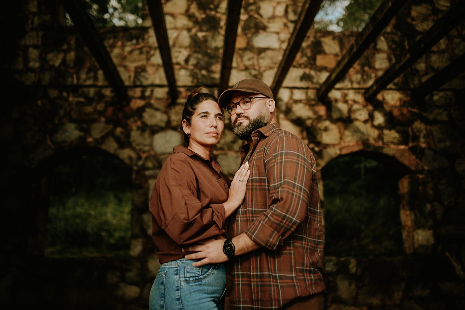 Couple in matching brown outfits embracing romantically under stone arches in a lush outdoor setting.