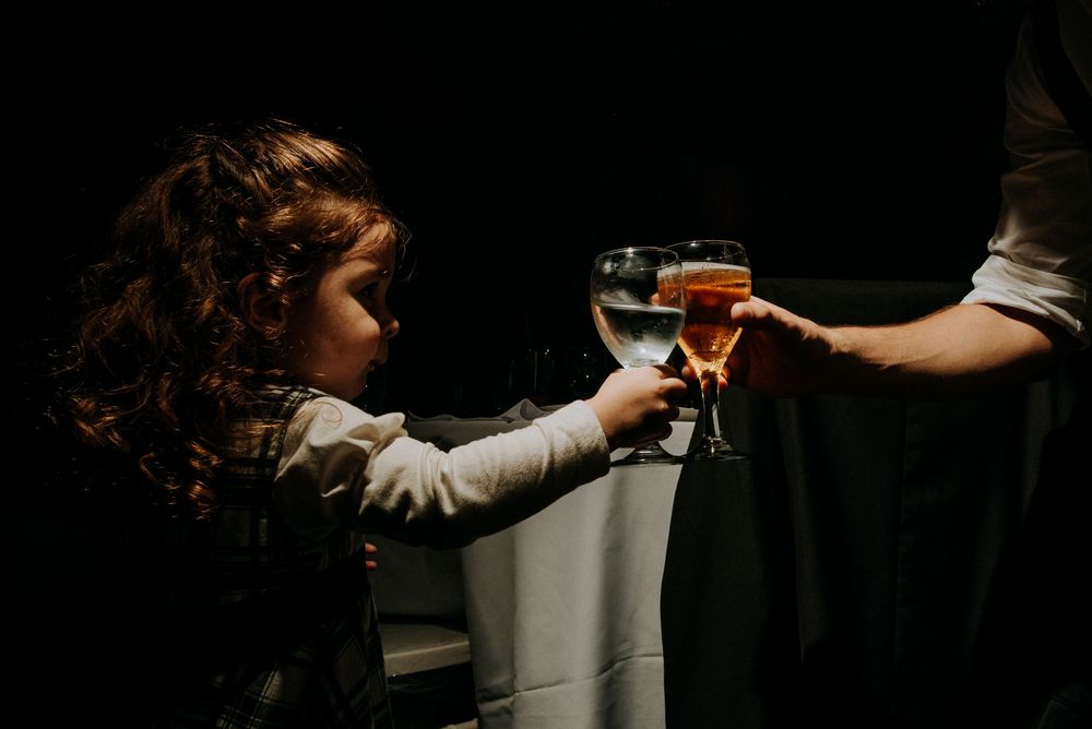 Young child clinking wine glasses with an adult in a dimly lit room at a formal event. Fotógrafo de bodas en Formosa