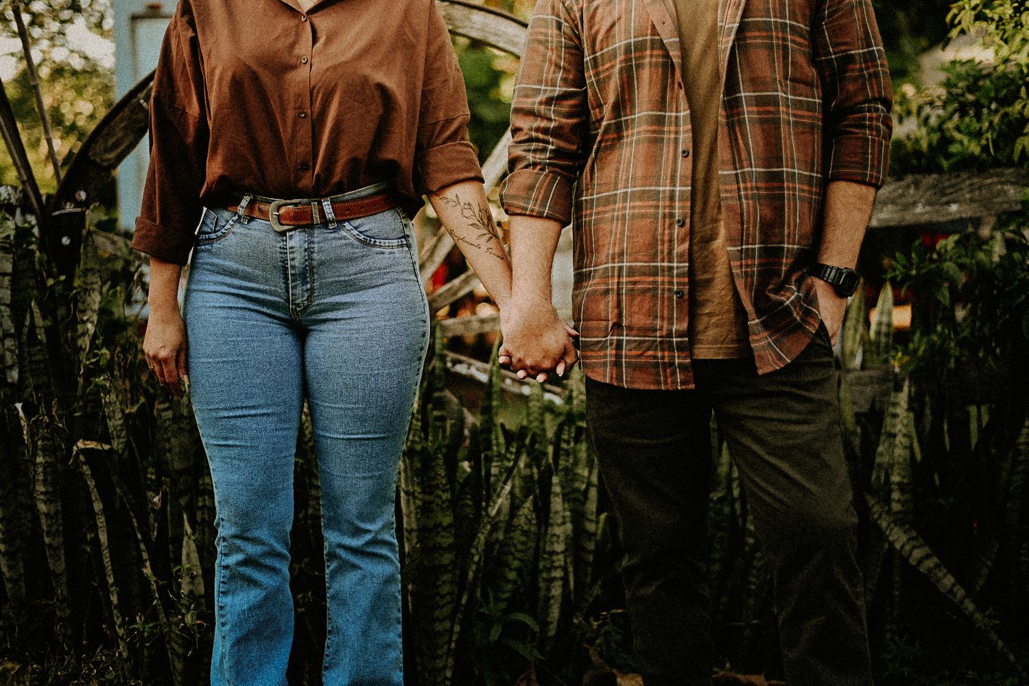 Couple holding hands outdoors in casual fall outfits, woman in jeans and man in plaid shirt, surrounded by lush greenery.