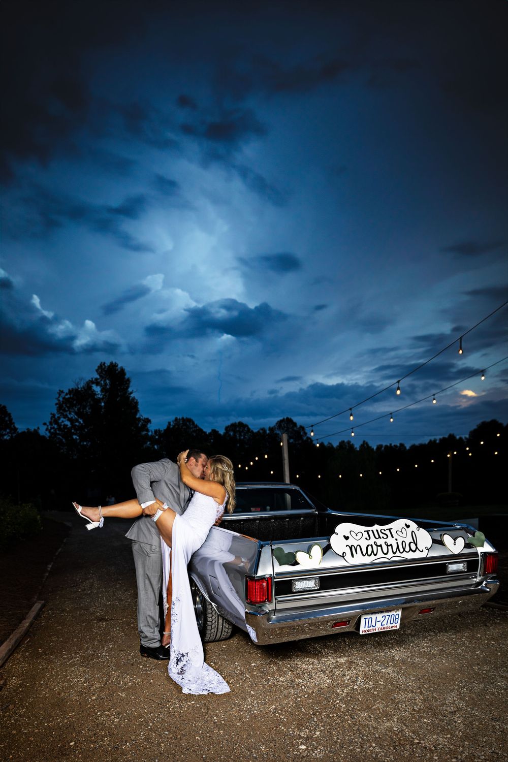 A romantic moment by a vintage car with 'Just Married' sign under dramatic evening sky.