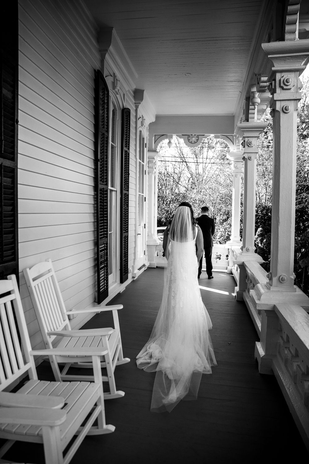 Black and white photo of a couple walking on a Southern-style porch with white rocking chairs and ornate columns.