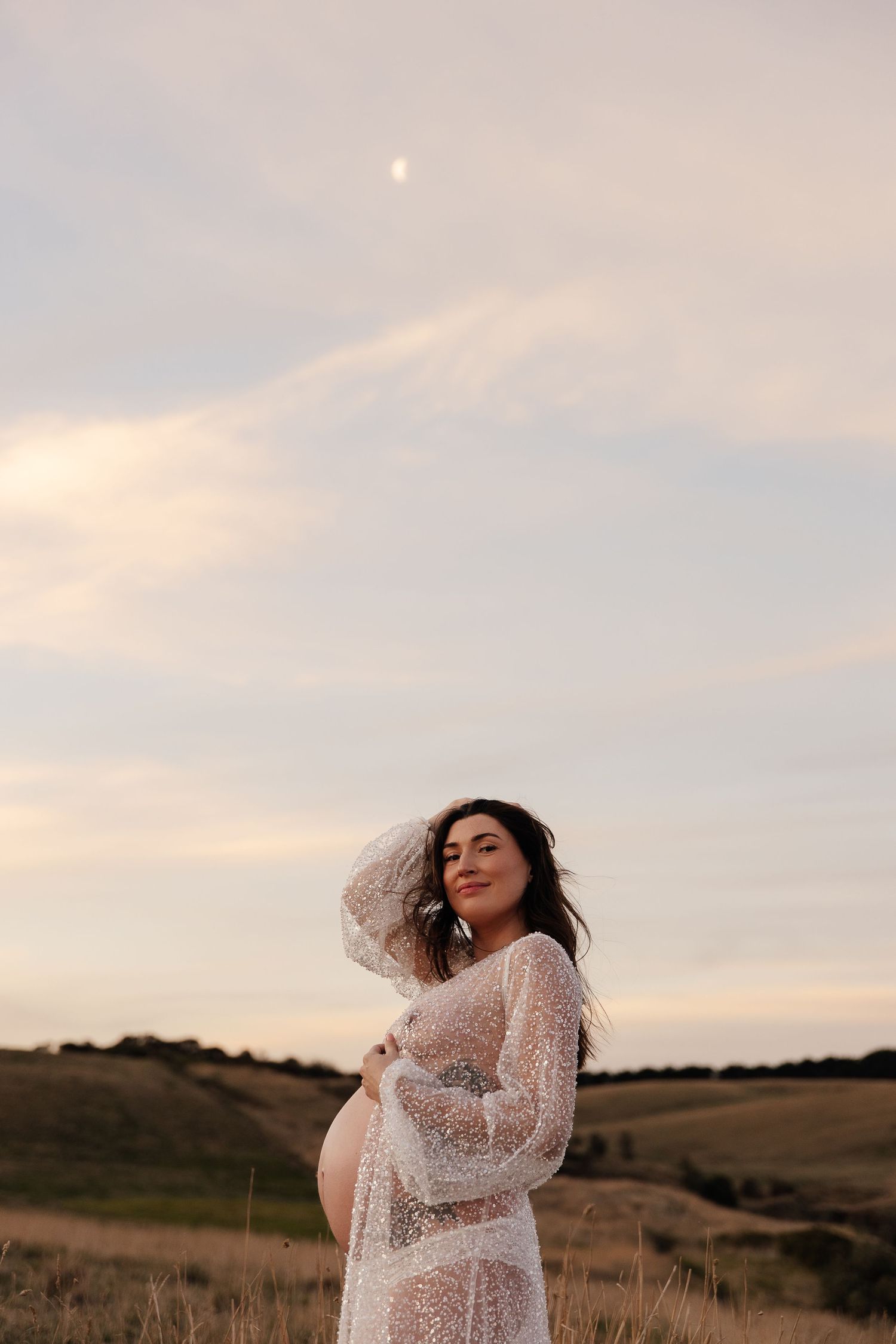 Pregnant woman in white dress poses joyfully on coastal cliffs at sunset.