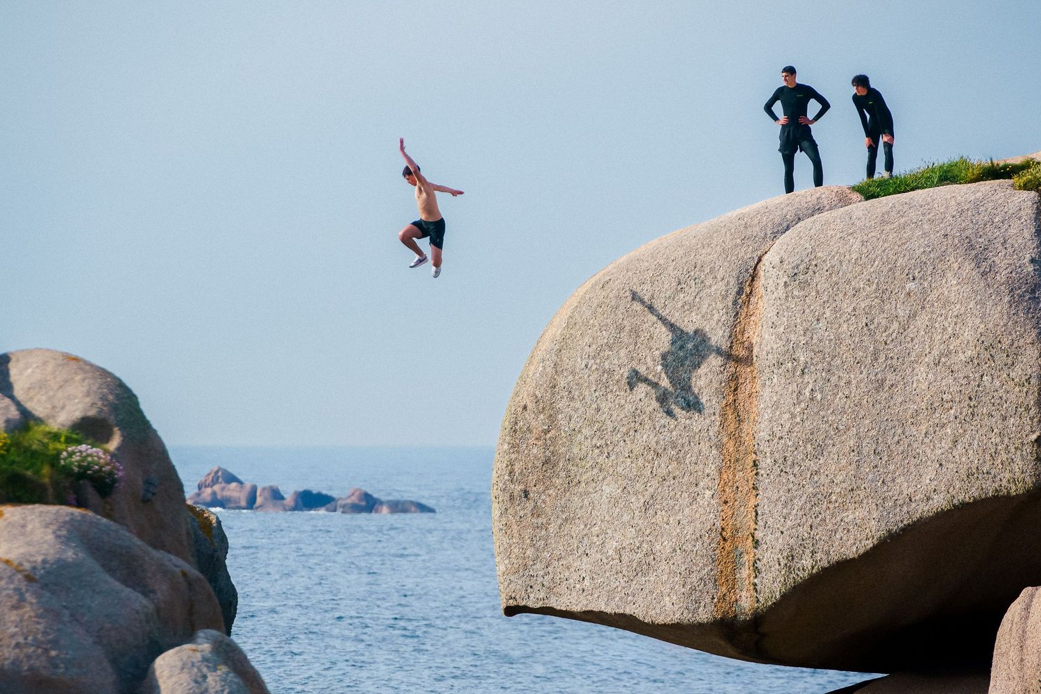 A daring cliff jumper leaps off a large granite rock into the ocean while two spectators watch from above.