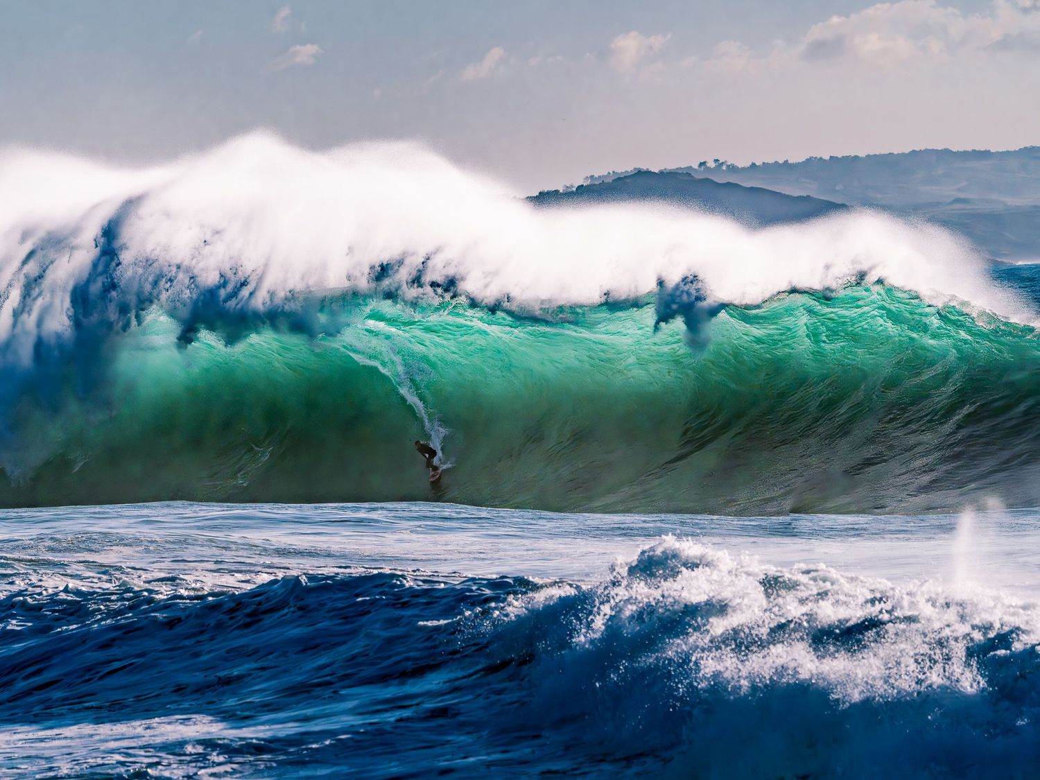 A surfer rides a massive turquoise ocean wave as it crashes with white foam against a hilly coastal backdrop.