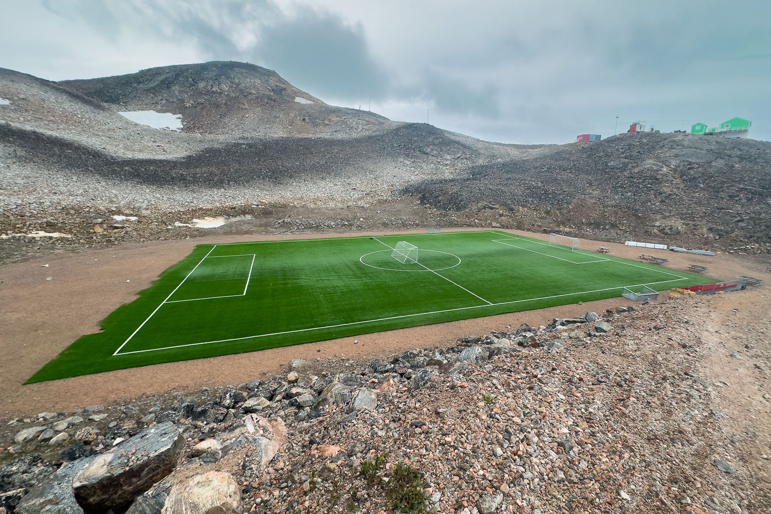 A pristine football pitch with bright green turf sits dramatically amid a rocky volcanic mountain landscape.