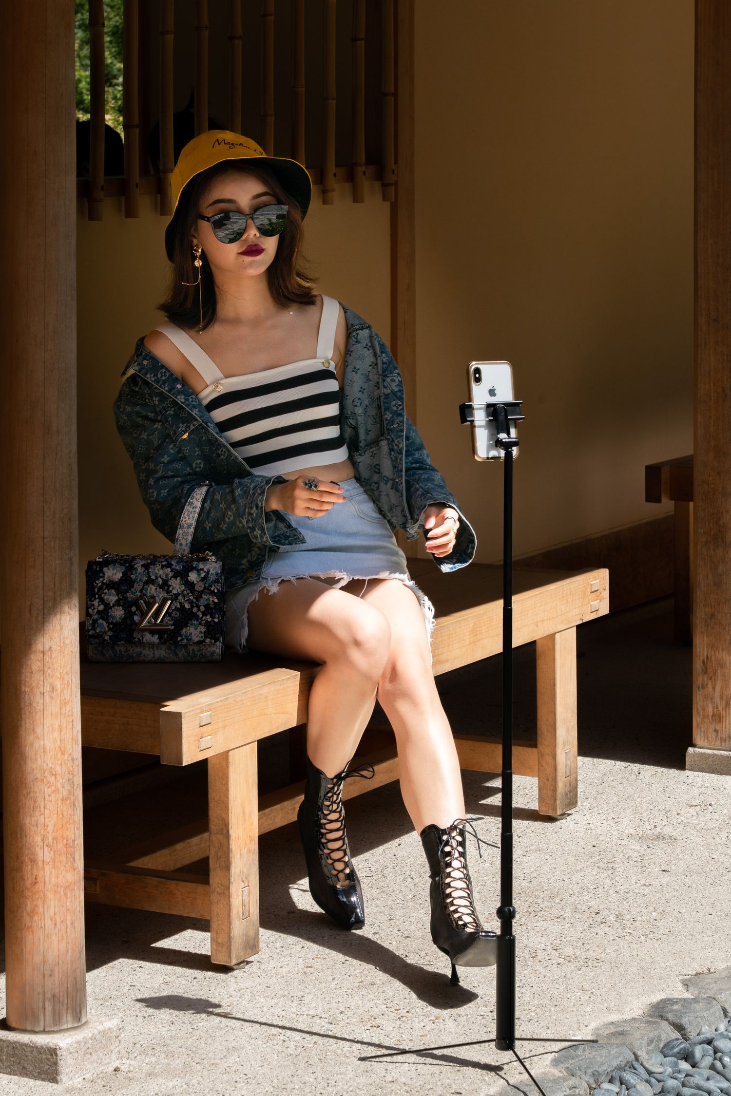 Stylish model in yellow hat, striped top and combat boots sitting on wooden bench taking selfie with phone on tripod.