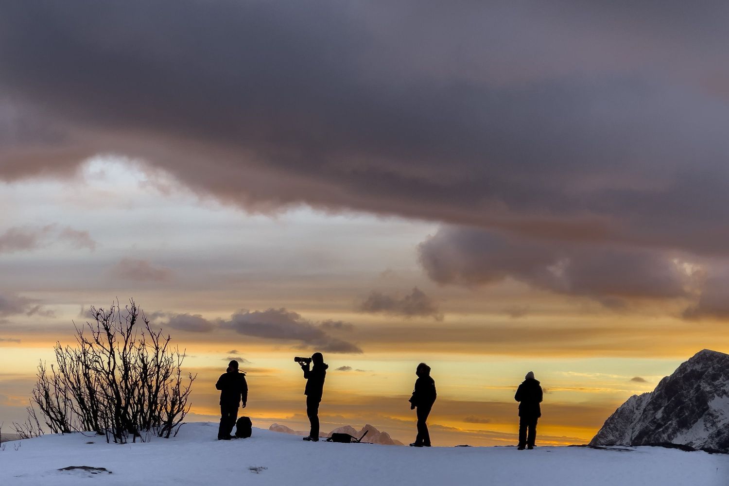 People stand in silhouette against a dramatic sunset sky with mountain peaks and orange clouds on the horizon.