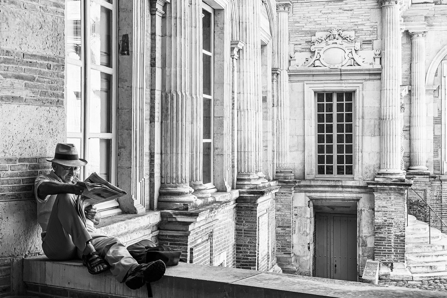 Someone in a hat sits reading on ornate classical architecture with columns and decorative stonework.