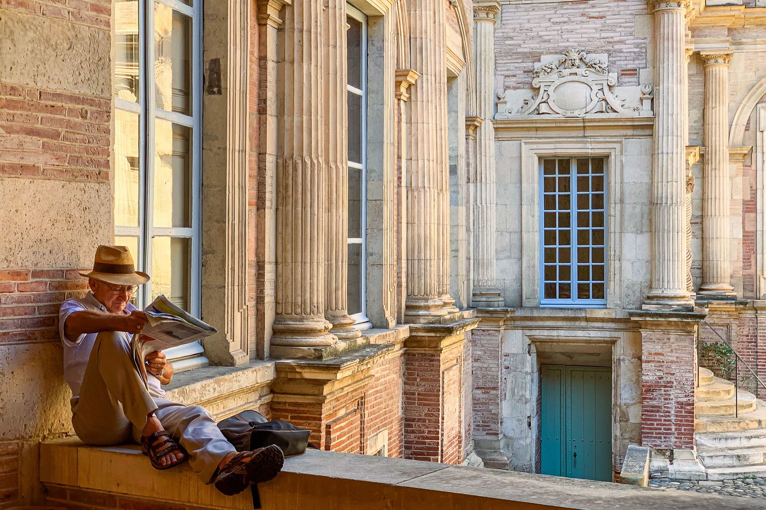 Elderly man reading on a ledge beside ornate classical columns of a historic brick building.