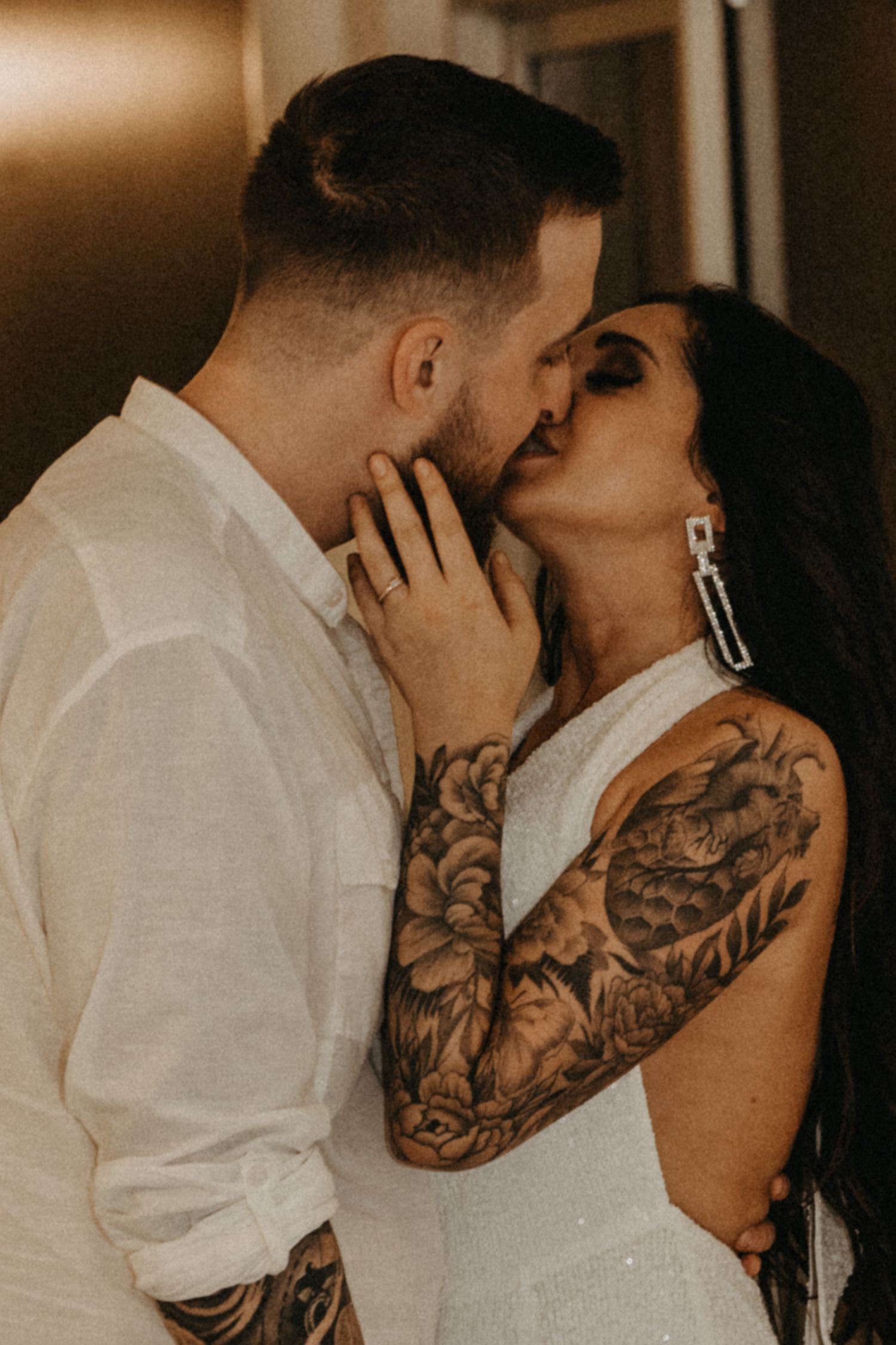 Tattooed couple sharing a passionate kiss, bride wearing white dress with sleeve tattoos visible.