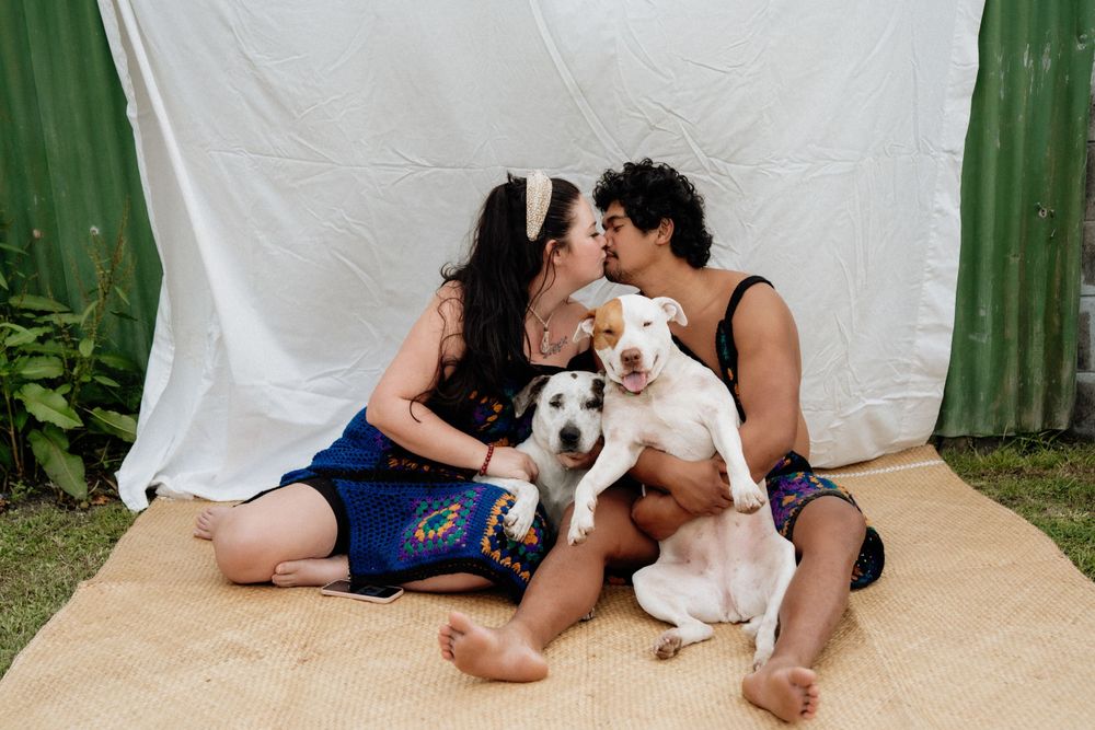 A couple sits on blanket outdoors with their white dog against white backdrop.
