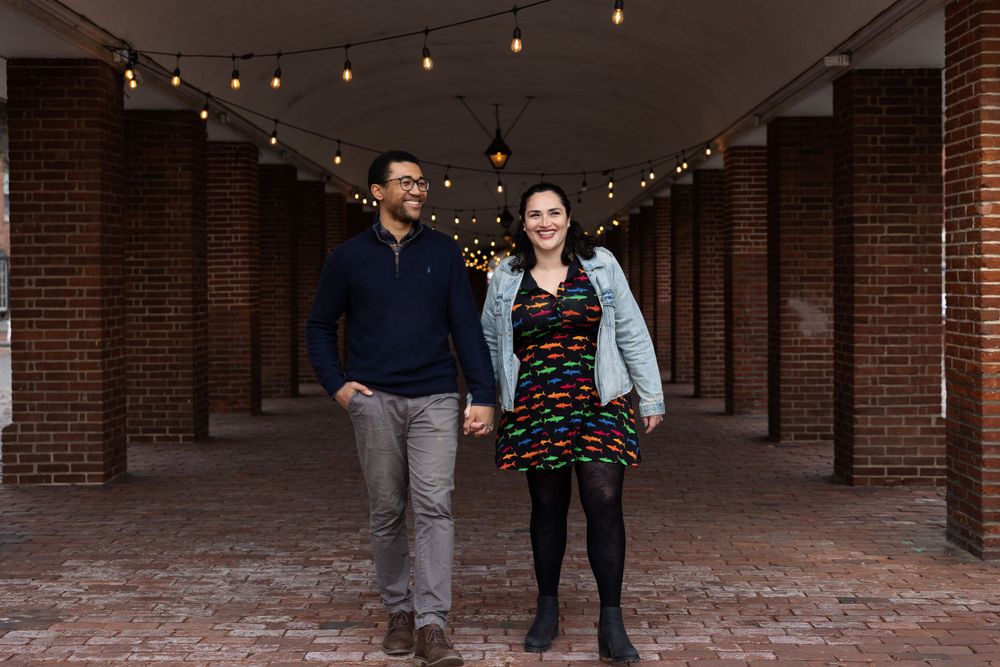 A couple walking hand in hand through a brick corridor with hanging lights.