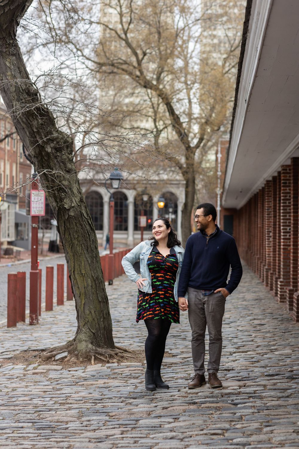 A couple strolls together on a historic cobblestone street with brick buildings and bare trees.