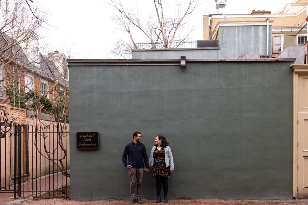 A couple poses against a grey wall outside a historic building.