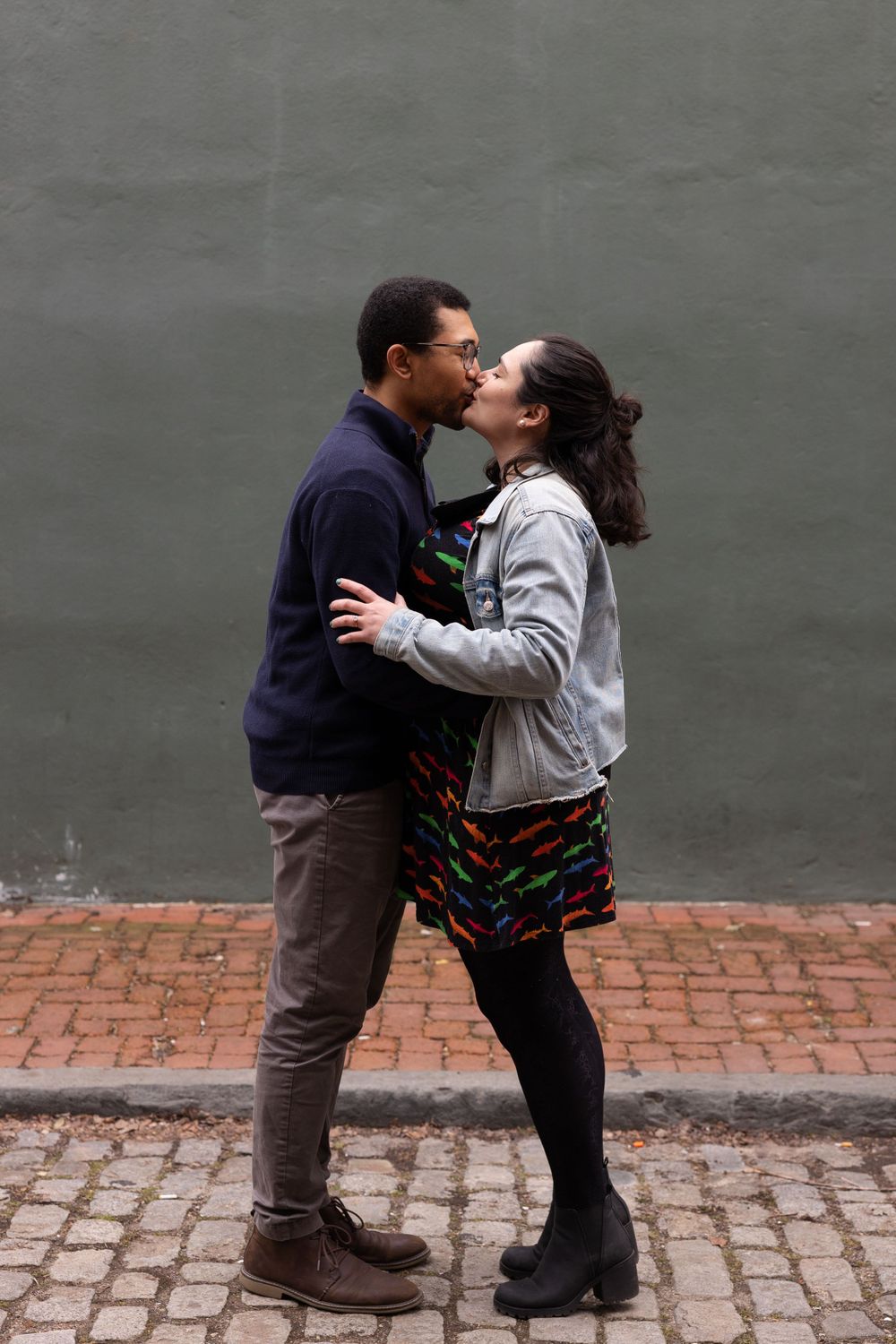 A romantic couple shares a kiss against a grey wall on a cobblestone street.