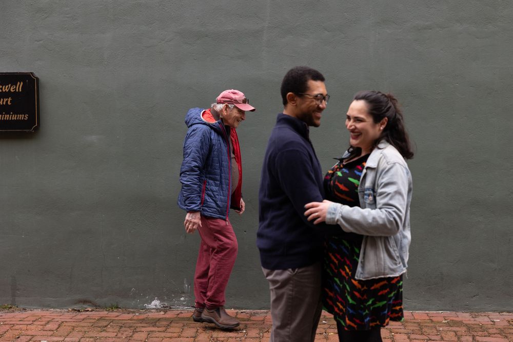 A couple shares a laugh while someone walks past in the background near a grey wall.
