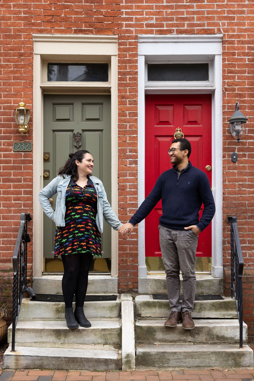 A couple stands on separate doorsteps between a grey and red door on a brick building.