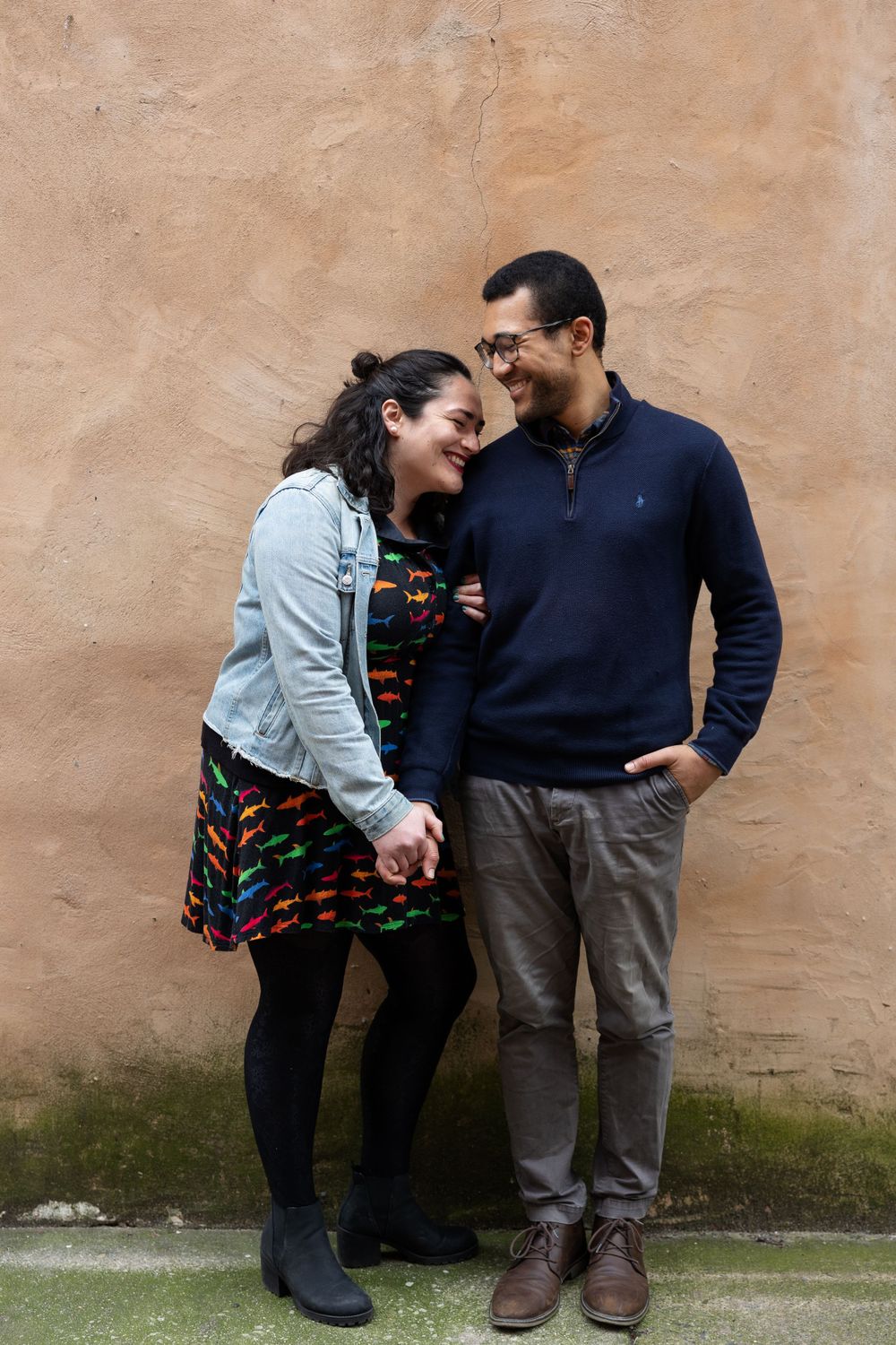 A couple leaning against a beige textured wall wearing casual outfits.