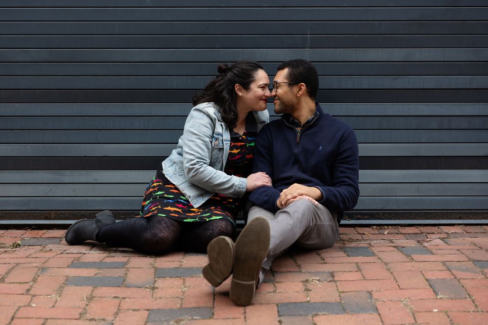 A couple sits intimately on brick pavement against a dark grey wall.