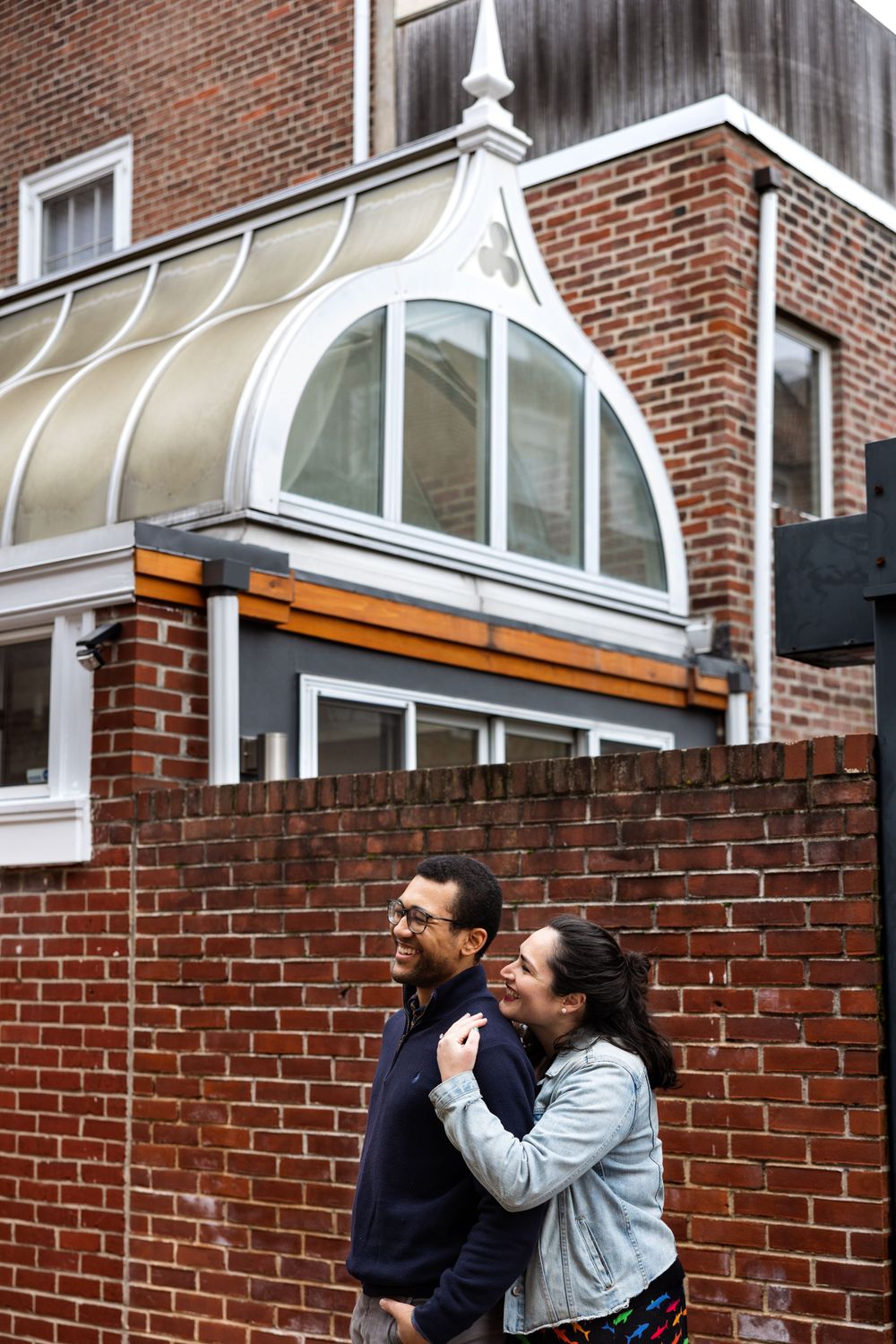 A couple embraces beneath an architectural glass dome feature on a brick building.