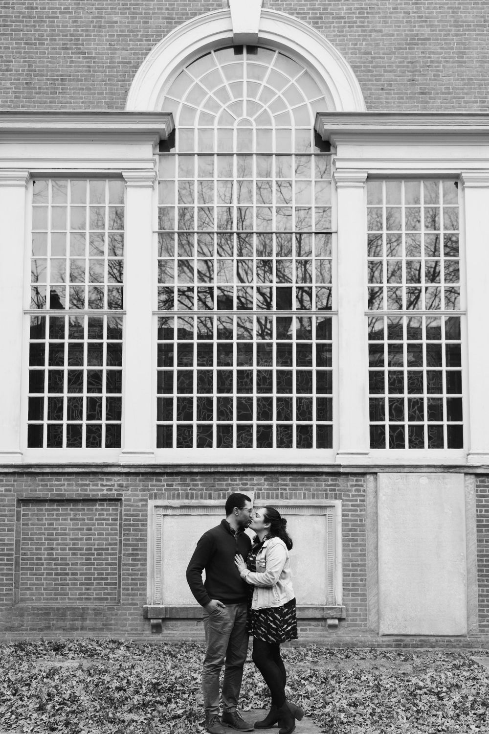 A couple embraces in front of a large arched window on a historic brick building.