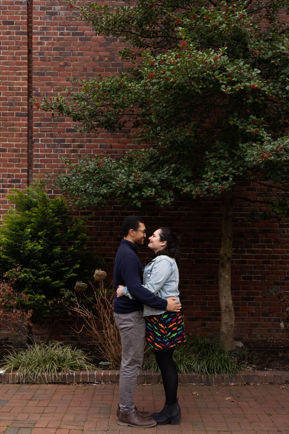 A couple embraces against a red brick wall with flowering shrubs.