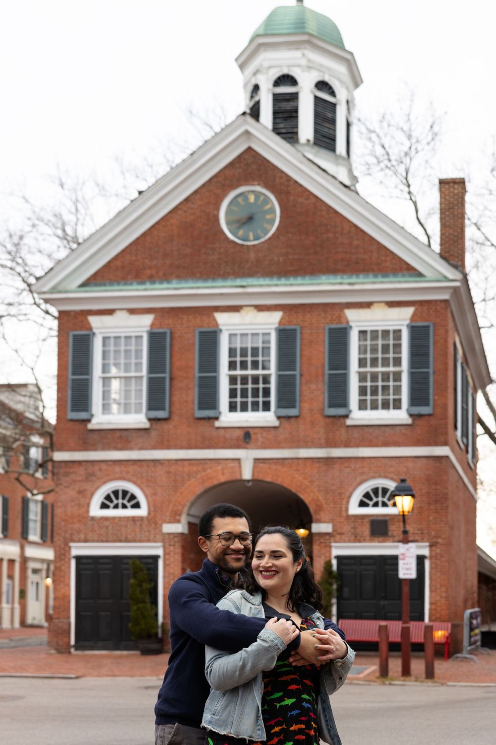 A couple poses playfully in front of a historic red brick building with green cupola.