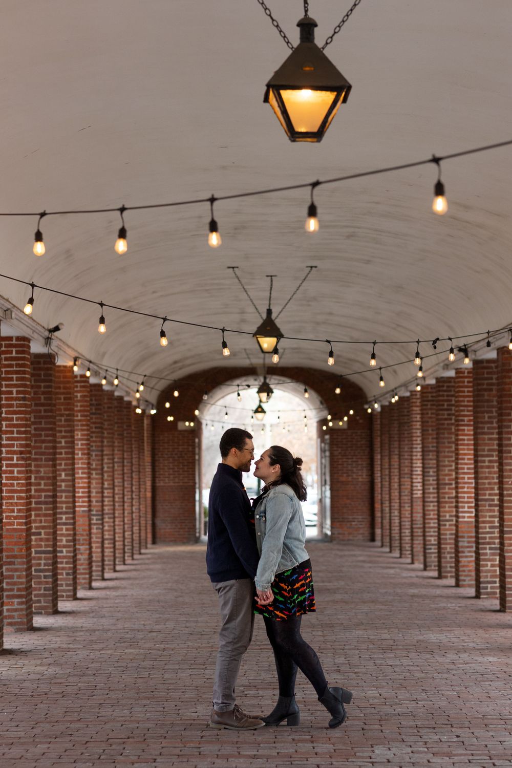 A couple stands in a brick archway tunnel with glowing string lights overhead.