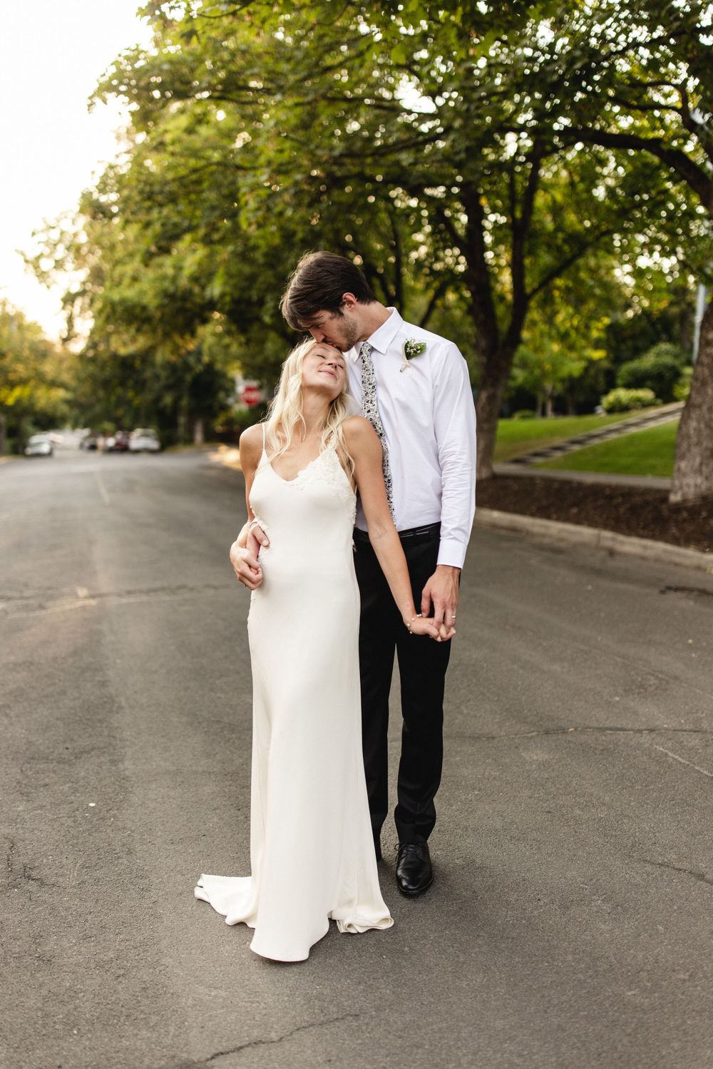 Groom tenderly kissing bride on the forehead while holding hands on a quiet tree-lined street at sunset.