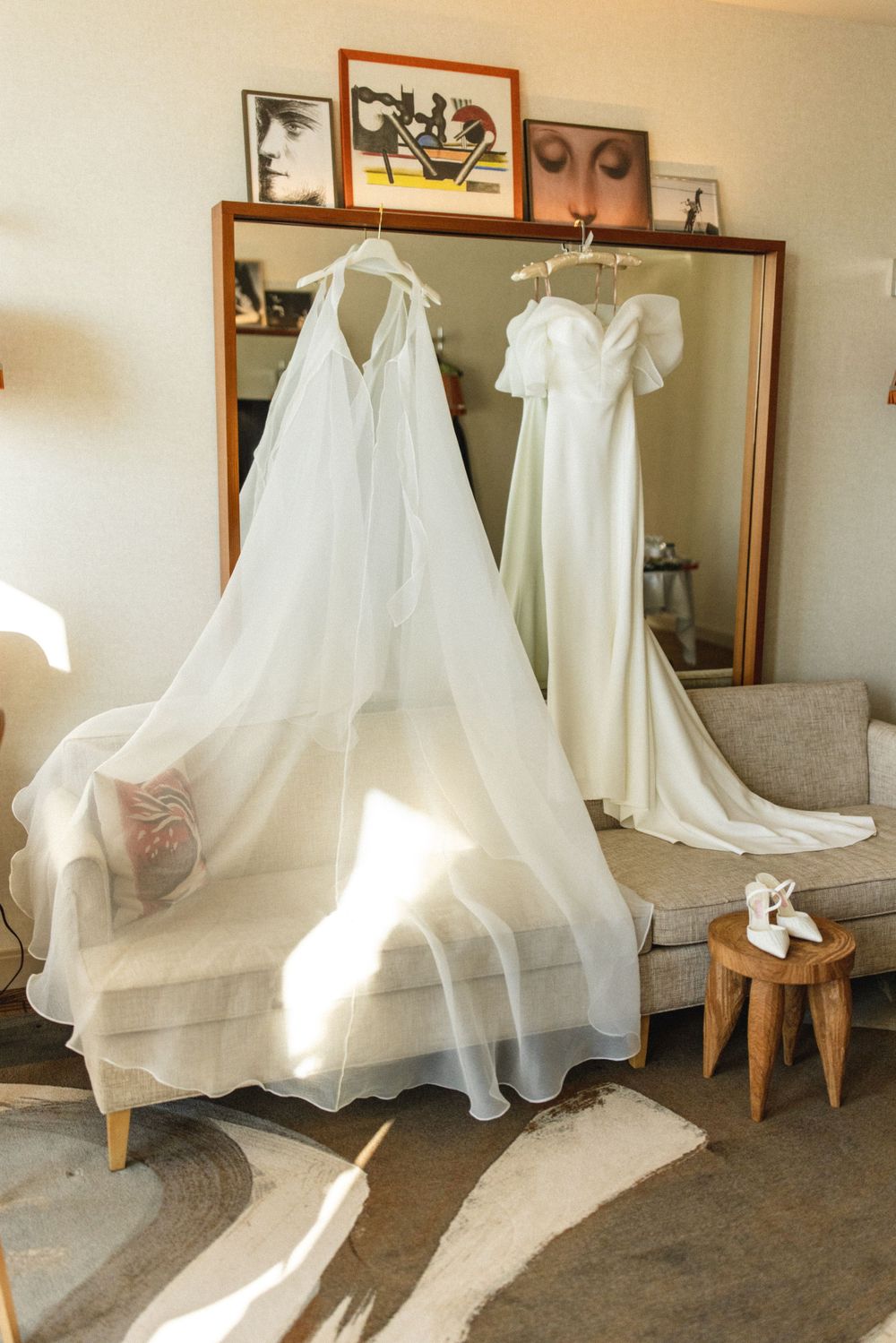 Two elegant white wedding gowns hanging on hangers reflected in a large mirror in a bright room.