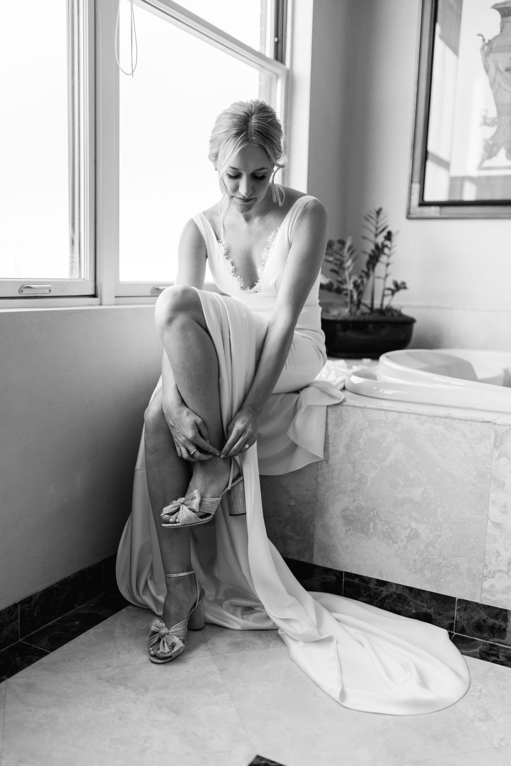 Black and white photo of blonde bride sitting on bathtub edge fastening wedding heels.