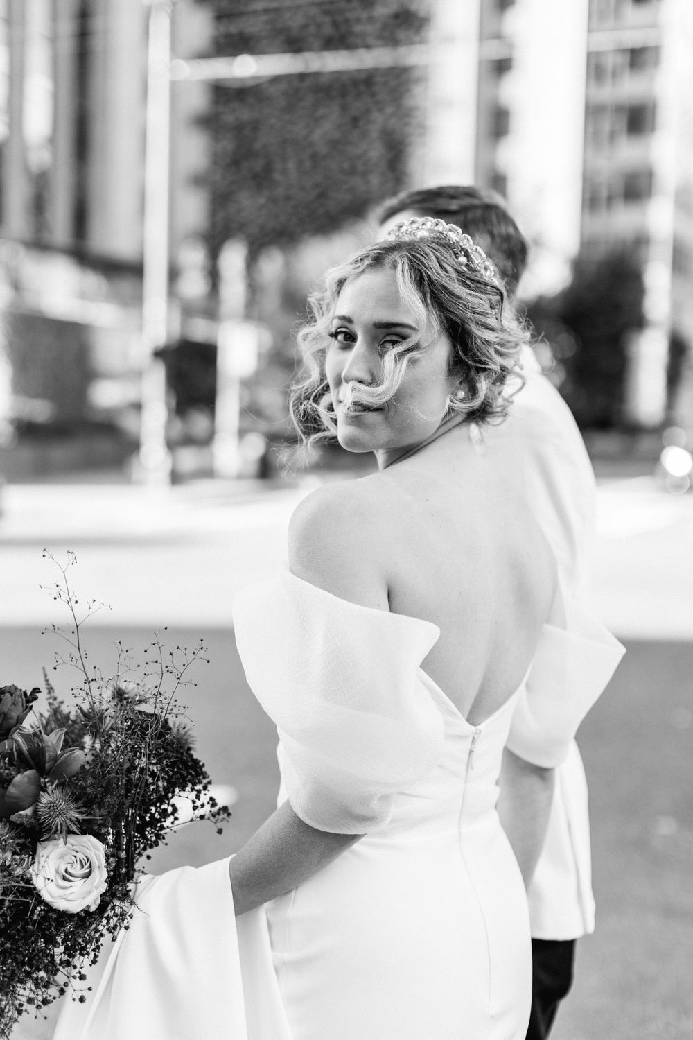 Black and white portrait of bride in off-shoulder gown holding bouquet on a city street.