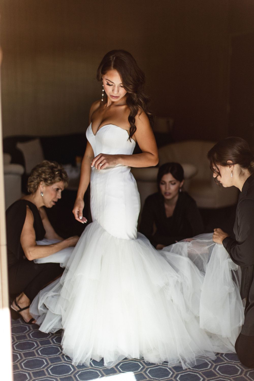 Bride in strapless mermaid wedding gown being helped by bridesmaids in hotel room before ceremony.