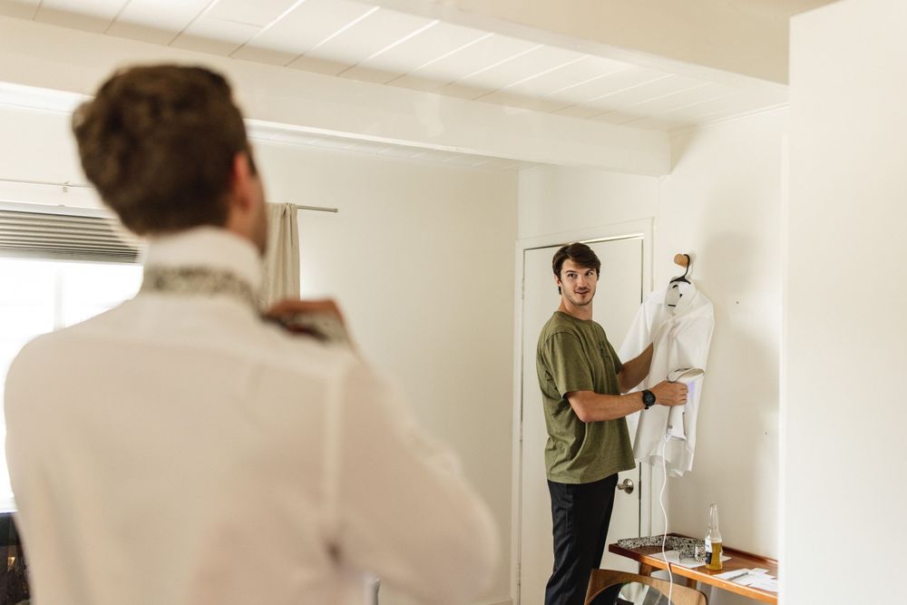 Groomsman in green shirt helping prepare a white wedding suit on a hanger in a bright white room.